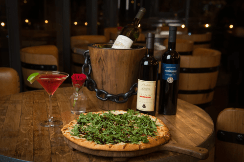 A wooden table with a pizza topped with arugula, a cocktail, and three bottles of wine in a bucket.