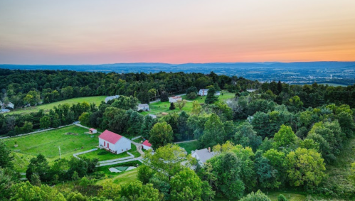 Aerial view of a lush green landscape with houses and a red-roofed barn at sunset, surrounded by rolling hills.