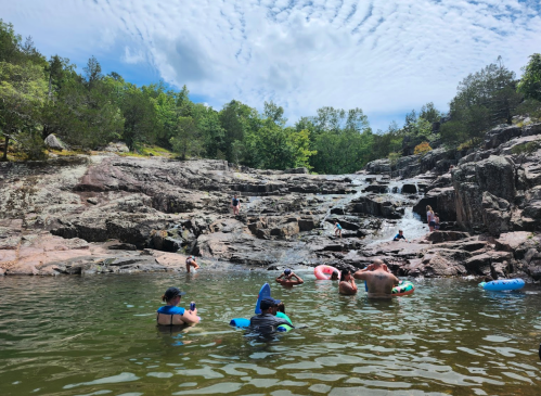 People swimming and relaxing in a natural pool surrounded by rocky terrain and lush greenery under a cloudy sky.