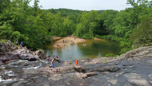 A scenic view of a natural swimming area surrounded by trees, with people enjoying the water and rocky terrain.