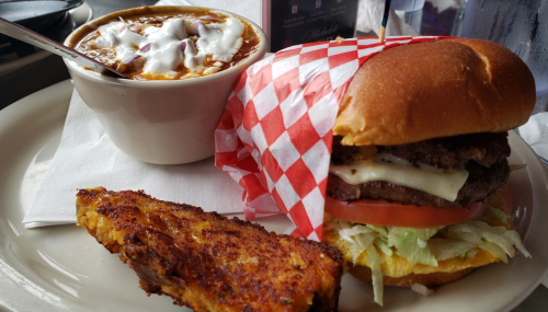 A plate featuring a burger with cheese, lettuce, and tomato, a bowl of chili, and a slice of cornbread.