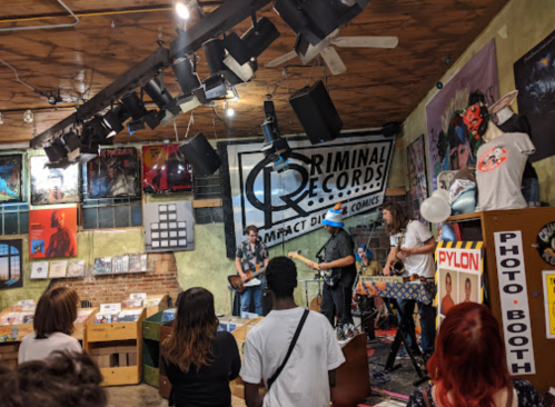 A small crowd watches a live band perform in a record store, surrounded by colorful album covers and merchandise.