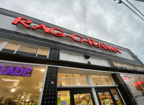 Storefront of Rag-O-Rama with bright red signage against a cloudy sky. Neon "Trade" sign visible in the window.