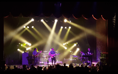 A live band performs on stage with colorful lights and a cheering audience in a concert setting.