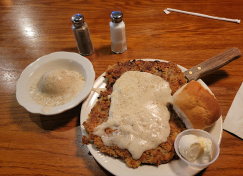 A plate of chicken-fried steak topped with gravy, a roll, and a side of mashed potatoes with butter, on a wooden table.
