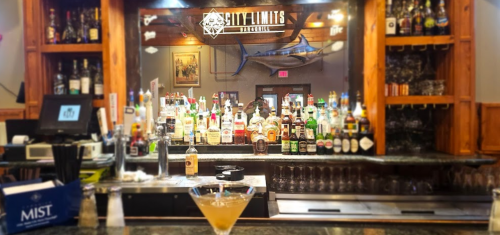 A bar with a variety of liquor bottles on shelves, featuring a cocktail in the foreground and a marlin mounted on the wall.