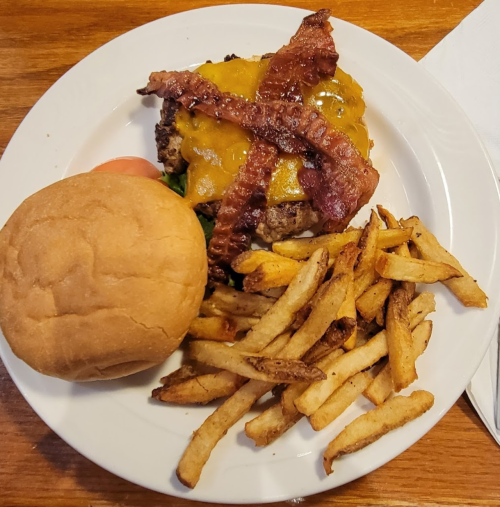 A plate with a cheeseburger topped with bacon, lettuce, and tomato, served with a side of golden French fries and a bun.