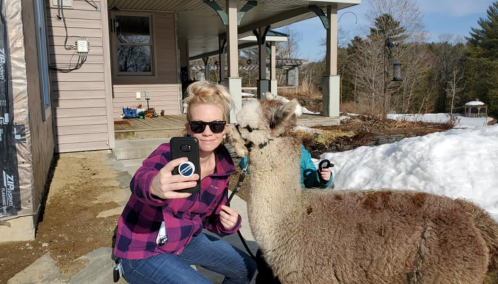 A woman in sunglasses takes a selfie with a llama outside a house, with snow on the ground and trees in the background.