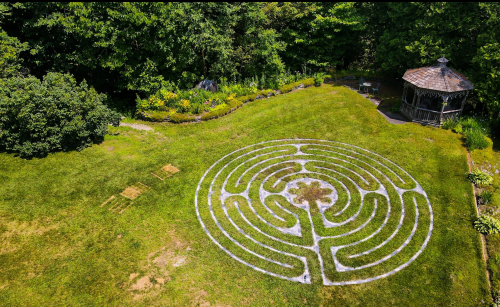 Aerial view of a circular maze in a grassy area, surrounded by trees and a gazebo in the background.