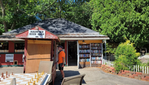 A quaint bookshop labeled "Haunted Book Shop" with outdoor seating and a chessboard in front, surrounded by greenery.