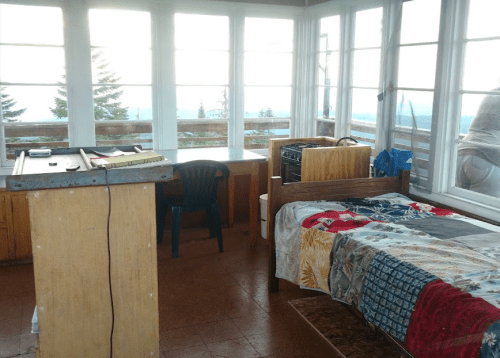 Cozy mountain lookout interior with a bed, desk, and large windows offering scenic views.