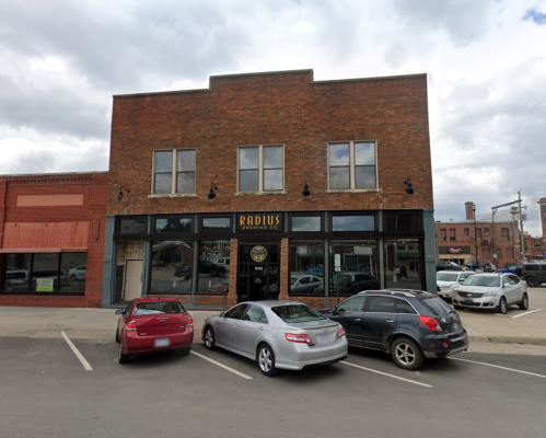A brick building with large windows and a sign reading "RADIUS," surrounded by parked cars on a street.