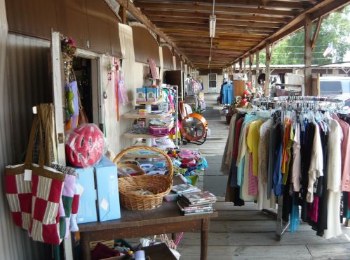 A thrift store aisle with clothing racks, shelves of items, and a fan, all under a wooden roof.