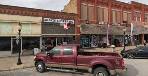 A red truck parked in front of Smokin' Sisters BBQ and nearby shops on a cloudy day.