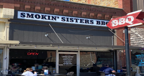 Exterior of Smokin' Sisters BBQ restaurant with an "Open" sign and outdoor seating. Bright, sunny day.