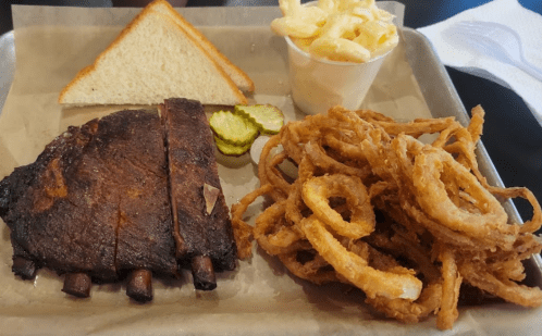 A plate with smoked ribs, pickles, onion rings, macaroni and cheese, and a slice of bread on parchment paper.