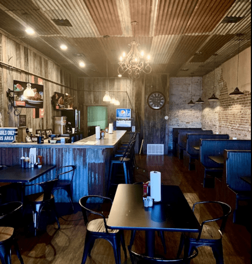 Interior of a rustic restaurant with wooden walls, a bar area, and black tables and chairs. Warm lighting creates a cozy atmosphere.