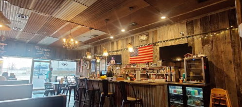 Cozy bar interior with wooden walls, an American flag, and a well-stocked counter, featuring warm lighting and seating.