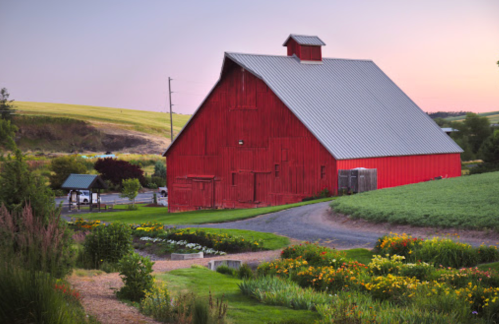 A red barn stands beside a garden, with rolling hills in the background under a pastel sky at dusk.