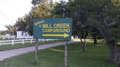 Sign for Mill Creek Campground with an arrow pointing right, surrounded by trees and a grassy area.
