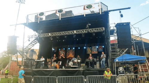 A live band performs on stage at an outdoor event, with a crowd and equipment visible in the background.