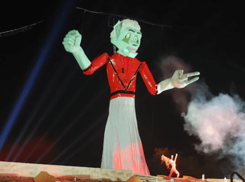 A large, illuminated effigy with a red outfit and green face, surrounded by smoke and lights at night.