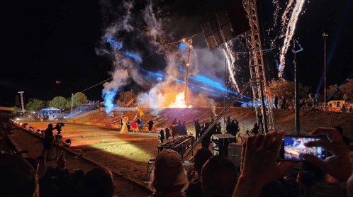 A nighttime event with fireworks and flames, featuring a crowd watching a performance on a lit stage.
