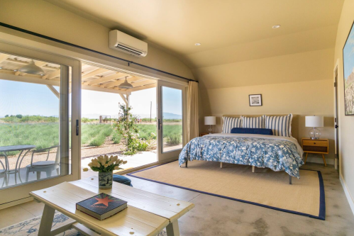 Cozy bedroom with a floral bedspread, large windows, and a view of greenery outside. Simple decor and natural light.
