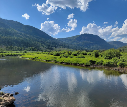 A serene landscape featuring a river reflecting mountains and clouds, surrounded by lush greenery under a blue sky.