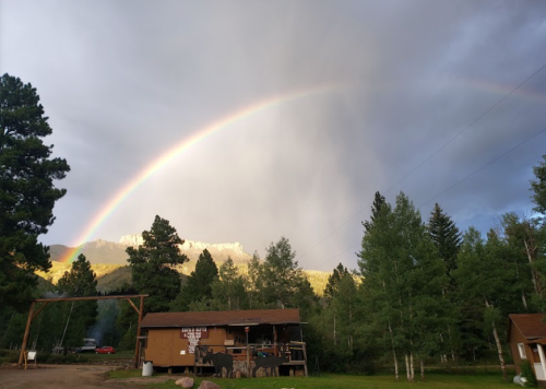 A rustic cabin surrounded by trees under a rainbow and cloudy sky, with mountains in the background.
