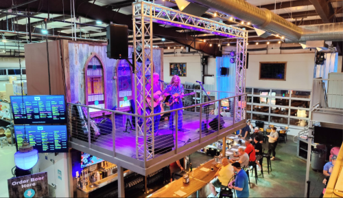A musician performs on a stage above a lively bar with patrons enjoying drinks and food below.