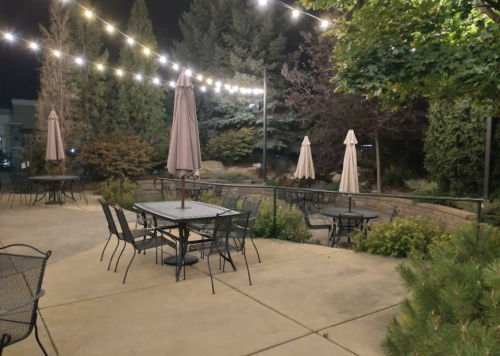 Outdoor patio at night with string lights, tables, and umbrellas surrounded by trees and greenery.
