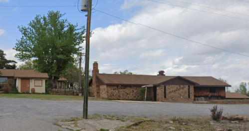 Two brick houses with a large tree nearby, set against a cloudy sky and a gravel road. A utility pole stands in the foreground.