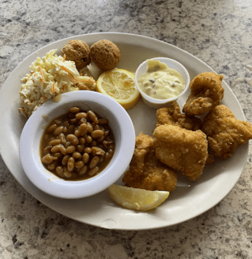 A plate of fried fish, coleslaw, baked beans, hushpuppies, lemon slices, and a small cup of tartar sauce.