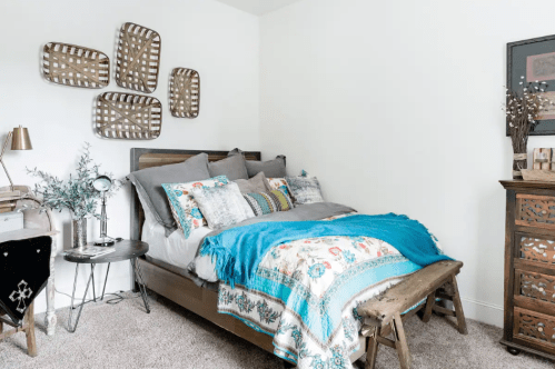 Cozy bedroom featuring a bed with decorative pillows, a blue throw, and woven wall art above a wooden bench.