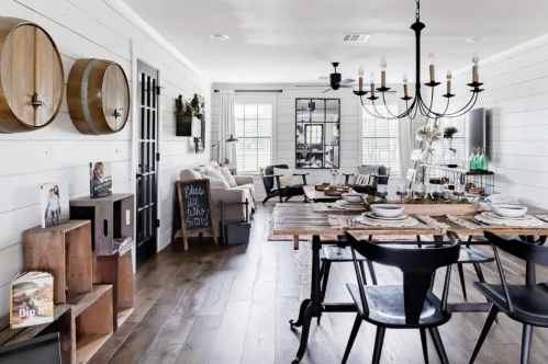 A cozy dining area with a wooden table, black chairs, and rustic decor, featuring a chandelier and large windows.