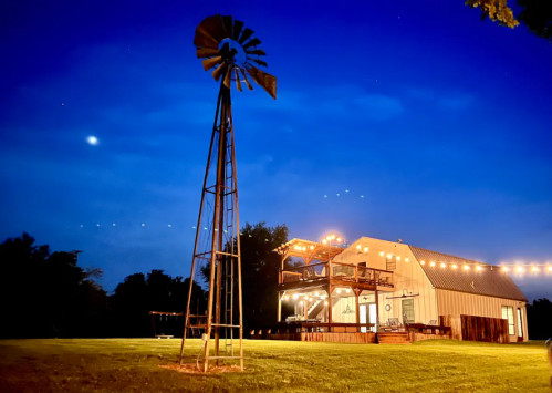 A rustic barn with string lights at dusk, featuring a windmill in the foreground and a clear blue sky.