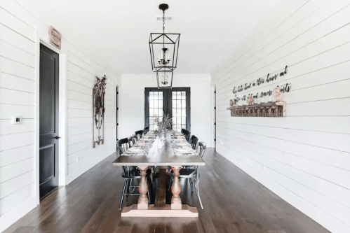 A spacious dining room with a long wooden table, black chairs, and white shiplap walls, featuring decorative wall art.