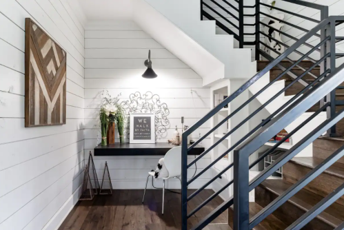 Modern entryway with a black desk, white walls, decorative art, and a staircase. A cat sits on a chair.