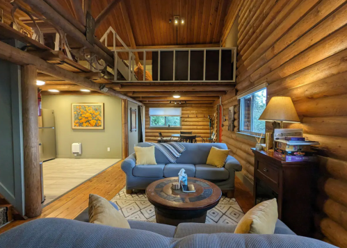Cozy log cabin interior with a living area, two sofas, a coffee table, and a lofted space above. Warm wooden accents.