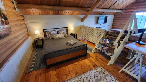 Cozy cabin interior featuring a bed, wooden beams, and a small seating area with a staircase. Warm, inviting atmosphere.