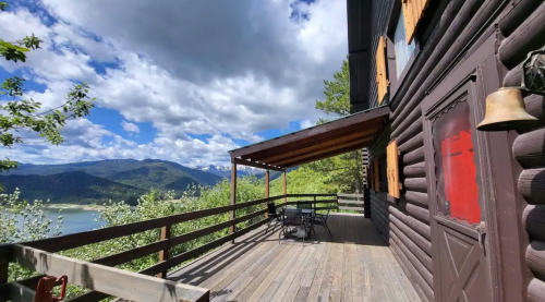 A wooden deck with a table and chairs overlooks a scenic mountain landscape under a partly cloudy sky.