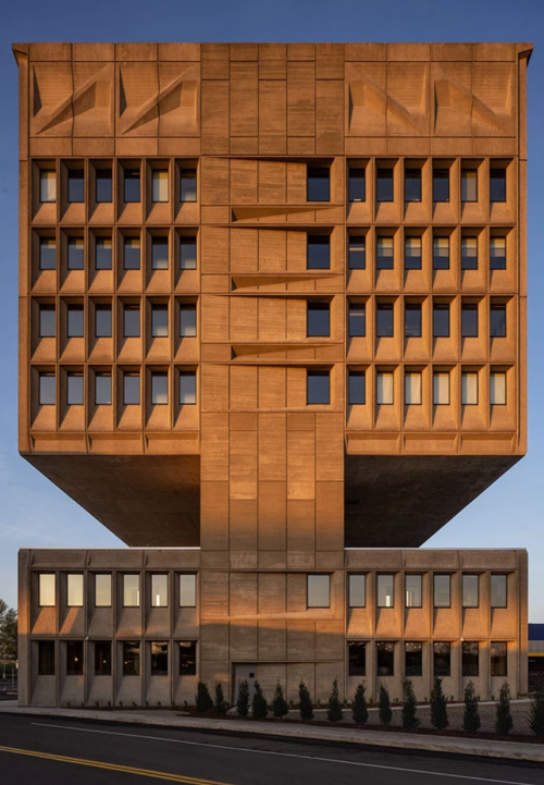 A modern, angular building with a textured facade, elevated on a concrete base, illuminated by warm sunlight.