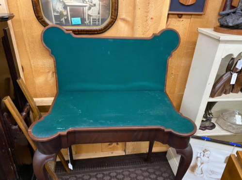 A vintage table with a green felt top and decorative wooden edges, set against a wooden wall in an antique shop.