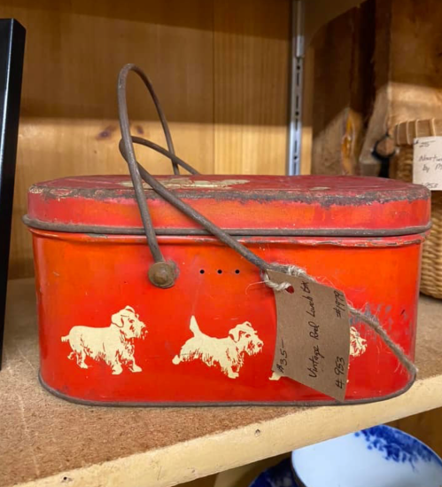 A vintage red tin box with a handle, featuring white dog silhouettes, displayed on a wooden shelf.