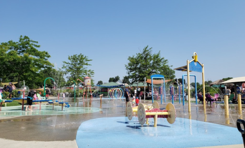 A vibrant splash pad with water features, surrounded by trees and families enjoying a sunny day.
