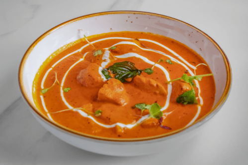 A bowl of creamy orange curry with chicken pieces, garnished with herbs and a swirl of cream.
