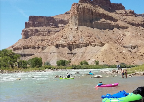 People kayaking in a river surrounded by rocky cliffs and greenery on a sunny day.