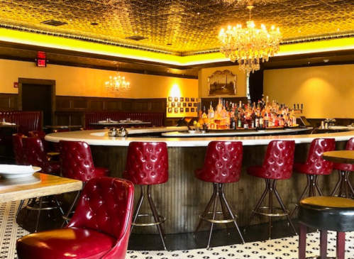 Elegant bar interior featuring red leather chairs, a circular bar, and a chandelier under a decorative ceiling.
