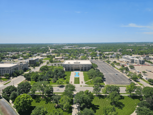 Aerial view of a green landscape with a fountain, buildings, and a clear blue sky in the background.
